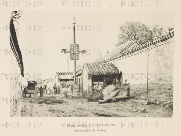 Street with customs posts in Beijing, Pékin. - La rue des customs, page, photomechanical print, inscription: 'Photographie de l'autoriteit.', photographer: Hugues Krafft, Beijing, cliché maker: anonymous, in or after 1882 - in or before 1883, Hugues Krafft, photographer, cliché maker, paper, collotype, photoprint, height 108 mm x width 166 mm, street, façade (of house or building), customs, A narrow dirt street lined with brick walls and low tiled-roof buildings, a tall ornate pole with decorative crossarms standing near the foreground, a small shop or house with an open window and stacked bundles or hay nearby, groups of people walking or standing along the street, a covered cart facing away from the viewer, tiled rooflines and trees visible above the walls, and a worn uneven pathway with scattered stones.