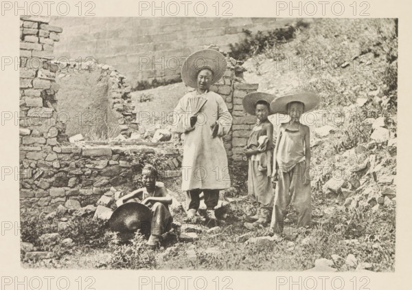 Tour guide Barthélemy and three mule drivers in front of a ruin in China, Barthélemy et les trois aniers, page, photomechanical print, inscription: 'Photographie de l'autoriteit.', inscription: 'HK', photographer: Hugues Krafft, China, cliché maker: anonymous, in or after 1882 - in or before 1883, Hugues Krafft, photographer, cliché maker, paper, collotype, photoprint, height 113 mm x width 168 mm, boy (child between toddler and youth), ruin of a building, architecture, fan, A black-and-white photograph showing four people standing and sitting among rough stone walls and a rocky, grassy hillside; one person stands near the center wearing a long light-colored garment and a wide straw hat while holding a folded hand fan, two younger individuals stand to the right wearing loose clothing and wide straw hats, and one person sits on the ground at the left foreground with a large round metal pan resting near their legs; broken stones, rubble, and patches of vegetation surround them and a part...