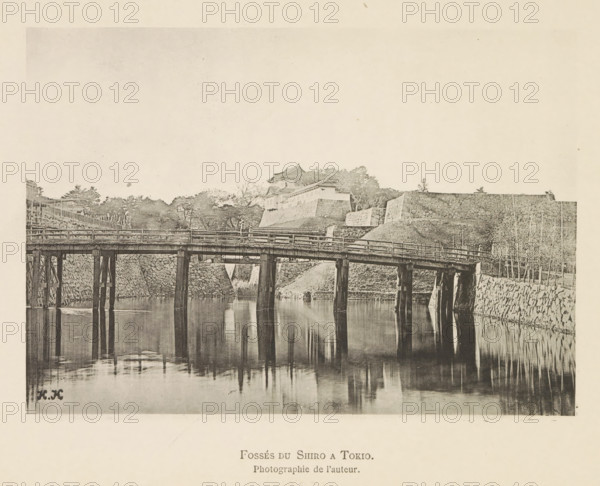 Bridge over a canal in Tokyo, with the Kokyo, Fossés du Shiro a Tokyo in the background, page, photomechanical print, inscription: 'HK', inscription: 'Photographie de l'autoriteit.', photographer: Hugues Krafft, Tokyo, cliché maker: anonymous, in or after 1882 - in or before 1883, Hugues Krafft, photographer, cliché maker, Kanto, paper, collotype, photoprint, height 115 mm x width 164 mm, bridge, palace, moat, fortified city, A sepia-toned photograph showing a wooden bridge supported by vertical posts spanning calm reflective water, with the bridge's railings and structural beams visible; behind the bridge are sloped stone embankments and terraced stone walls leading up to a cluster of traditional roofed buildings partially obscured by trees; bare and leafy trees line the top of the embankments and small poles or stakes stand along the right side near the water's edge; the water surface reflects the bridge posts and nearby stonework, and the sky above is light and largely empty.