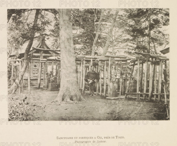 View of an Oji shrine near Tokyo, Sanctuaire et portiques a Oji, près de Tokyo, page, photomechanical print, inscription: 'Photographie de l'autoriteit.', photographer: Hugues Krafft, Japan, cliché maker: anonymous, in or after 1882 - in or before 1883, Hugues Krafft, photographer, cliché maker, paper, collotype, photoprint, height 117 mm x width 170 mm, forest, wood, road side shrine, Kanto, Tokyo, A sepia-toned scene shows a fenced wooden structure with vertical posts and horizontal beams set among tall trees, a large tree trunk occupying the foreground center with visible roots at its base, a person standing near the fence wearing traditional clothing and a head covering, a small stone statue on a pedestal beside the fence, a building with a sloped roof partially visible behind the fence and trees, scattered leaves and bare ground in the open area in front, and branches and foliage filling the upper portion of the image.