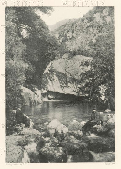 La Pentica, Two men before the Pentica Gorge in Corsica, page, photomechanical print, photographer: Roland Napoleon Bonaparte, Corsica, cliché maker: Lemercier & Cie., Paris, c. 1881 - in or before 1890, paper, photoaquatint, height 167 mm x width 118 mm, sitting figure, ravine, chasm, abyss, canyon, A grayscale scene shows a rocky river with smooth boulders in the foreground and a calm pool further back, flanked by steep rock faces and dense trees; a large rounded rock overhang sits above the pool with water flowing beneath it, and two men in dark coats and hats are seated on separate rocks near the water’s edge.