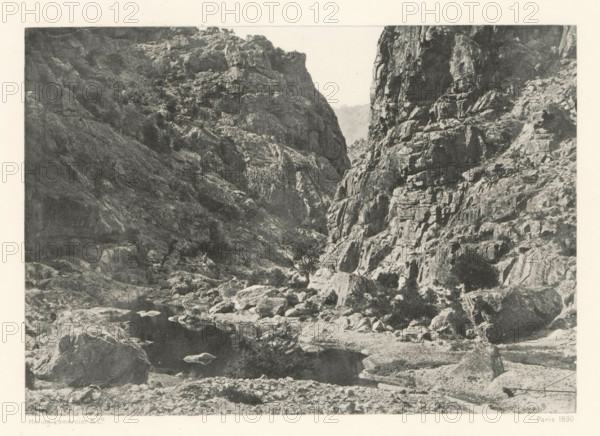 Gorges du Golo, Gorge of the river Golo in Corsica, page, photomechanical print, photographer: Roland Napoleon Bonaparte, Corsica, cliché maker: Lemercier & Cie., Paris, c. 1881 - in or before 1890, paper, photoaquatint, height 117 mm x width 167 mm, river, ravine, chasm, abyss, canyon, A monochrome image shows a rocky canyon with steep, jagged cliff faces on both sides and a narrow gorge between them. The foreground contains scattered boulders and a rocky shoreline leading to a dark, calm pool of water reflecting nearby stones. The cliff surfaces display rough textures, ledges, and crevices, with some sparse low vegetation clinging to the rock. The sky visible above the gorge is pale and contrasts with the darker tones of the rock.