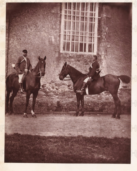 Two hunters on horseback, page, photomechanical print, photographer: Léon Cremière, France, cliché maker: anonymous, c. 1869 - in or before 1879, paper, Woodburytype, Woodburytypepie, height 202 mm x width 160 mm, Print on frontispiece., riding a horse, ass, or mule, rider, horseman, hunter, A sepia-toned photograph showing two men seated on horses in front of a textured stone wall with a tall window featuring a grid of vertical and horizontal bars; the riders wear jackets and breeches and hold reins, one rider facing slightly toward the camera while the other sits in profile, a coiled rope or lanyard visible over one rider’s shoulder, the horses are dark in color with bridles and saddles, the ground beneath them is a mix of dirt and a strip of grass in the foreground.