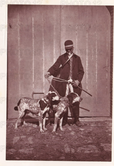 Hunter with the hunting dogs Cruelle and Généraux, page, photomechanical print, photographer: Léon Cremière, France, cliché maker: anonymous, c. 1869 - in or before 1879, paper, Woodburytype, Woodburytypepie, height 195 mm x width 132 mm, hunter, hunting dogs, A sepia-toned photograph showing a person standing in front of a tall wooden wall with vertical planks, wearing a dark coat, light trousers, gloves, and a cap with a horizontal band; the person holds a stick in one hand and several leashes or ropes in the other. In front of the person are two medium-to-large short-haired dogs with spotted coats and collars, standing close together and facing slightly to the right. The ground in the foreground is uneven and rocky. The image has a narrow white border around it.