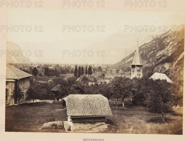 View of Meiringen, Meyringen, photograph, page, photographer: Francis Frith, Meiringen, c. 1855 - in or before 1865, paper, albumen print, height 120 mm x width 170 mm, city-view, and landscape with man-made constructions, parts of church exterior and annexes: tower, A sepia-toned landscape showing a village set against rolling hills and steep rocky mountains, with a tall church tower topped by a pointed spire and a weather vane visible near the right side, clustered buildings with pitched roofs and a larger multi-story building on the left, tree-lined fields and rows of trees stretching into the distance, fenced yards and scattered trees in the foreground, and a small wooden structure with a shingled roof positioned near the center foreground.