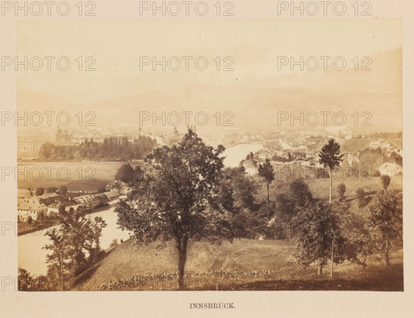 Innsbrück, View of Innsbruck, photograph, page, photographer: Francis Frith, Innsbruck, c. 1855 - in or before 1865, Tirol, paper, albumen print, height 121 mm x width 171 mm, river, city-view, and landscape with man-made constructions, A sepia-toned panoramic view from an elevated grassy hillside showing a large leafy tree in the foreground and additional trees scattered on the slope, with a river curving through a town of clustered buildings and rooftops beyond; the town includes rows of houses, larger structures with domed and peaked roofs, and a tree-lined riverbank, while distant hills rise faintly in the hazy background under a pale sky.