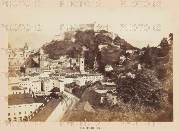 Saltzburg, View of Salzburg, photograph, page, photographer: Francis Frith, Salzburg (city), c. 1855 - in or before 1865, paper, albumen print, height 116 mm x width 166 mm, city-view, and landscape with man-made constructions, castle, A sepia-toned panoramic view of a historic town with clustered rooftops, narrow streets, and a mix of multi-story buildings and churches; several towers with domed and spired tops rise above the roofs, and a large fortress sits atop a wooded hill overlooking the town; the hillside shows terraced walls and scattered houses, while trees and foliage frame parts of the scene; in the foreground a long roofed structure and a curved street lead toward the denser center of buildings.