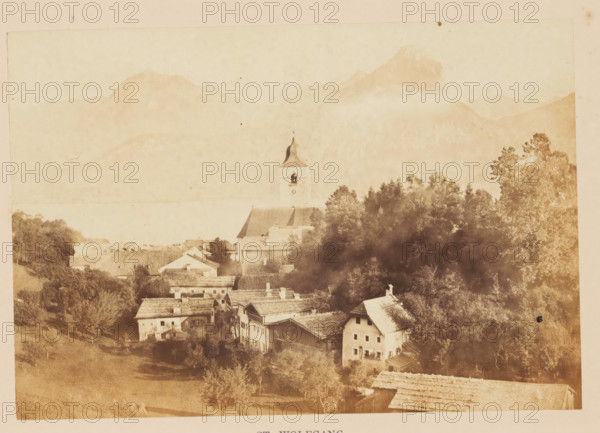 View of Sankt Wolfgang im Salzkammergut, St. Wolfgang, photograph, page, photographer: Francis Frith, Sankt Wolfgang, c. 1855 - in or before 1865, paper, albumen print, height 115 mm x width 168 mm, city-view, and landscape with man-made constructions, parts of church exterior and annexes: tower, mountains, A faded sepia view of a village with clustered buildings and sloped rooftops nestled among dense trees, a church tower with a pointed roof rising above the treeline, rolling hills and distant mountains forming a hazy backdrop, and open grassy areas in the foreground.