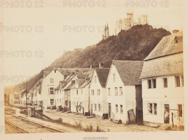 View of Stolzenfels Castle, presumably from Koblenz, Stolzenfels, photograph, page, photographer: Francis Frith, Koblenz, c. 1855 - in or before 1865, Rhineland-Palatinate, paper, albumen print, height 123 mm x width 174 mm, railway, tramway, rack railway, castle, Stolzenfels Castle, A row of light-colored, steep-roofed houses lines a street beside railway tracks, with some houses showing shuttered and open windows and small doors, a few people standing near the doorways and a figure near a window with hanging laundry; in the background a wooded hillside rises to a medieval-style castle with towers and spires perched atop the hill, and the scene has a sepia-toned, aged photographic appearance.
