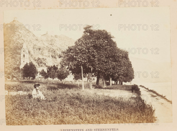 View of the bank of the Rhine and in the distance presumably Schloss Liebenstein, Liebenstein and Sternenfels, photograph, page, photographer: Francis Frith, Baden-Württemberg, c. 1855 - in or before 1865, paper, albumen print, albumendruk, height 116 mm x width 167 mm, castle, river bank, A sepia-toned scene shows a grassy foreground with a person seated wearing a wide-brimmed hat and light clothing, positioned near the lower left. A row of leafy trees with visible trunks lines the middle ground, casting darker foliage against a pale sky. Behind the trees on the left, a tall building with a pointed tower and arched openings stands at the base of a rocky hillside, which rises steeply and shows rough textures and outcrops. A narrow dirt path runs along the right edge toward a lighter area at the horizon.