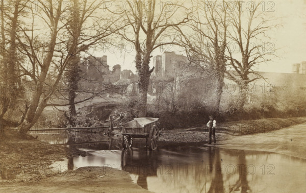 View of Kenilworth Castle, with a man with a handcart in the foreground, Kenilworth Castle, from the Brook, photograph, page, photographer: Stephen Thompson, c. 1854 - in or before 1864, Kenilworth, paper, albumen print, height 102 mm x width 162 mm, Photo opposite title page., hand-cart, ruin of a dwelling, house, castle, A sepia photograph showing a shallow river with a wooden cart partially in the water near the bank and its reflection visible on the river surface, several tall leafless trees with bare branches framing the scene, dense undergrowth and shrubs behind the trees, a simple wooden footbridge crossing the river in the midground, ruins of a large stone structure visible beyond the trees, a man standing on the near bank wearing a cap and vest and looking toward the camera, and soft reflections of trees and sky in the calm water.