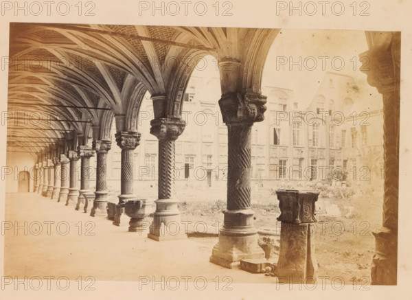 Court of the bishops palace, Liege, photograph, page, Liège, c. 1856 - in or before 1866, photographer, paper, albumen print, height 120 mm x width 183 mm, arcade, architecture, Palace of the Prince-Bishops, A long covered arcade with a series of stone columns and carved capitals supporting ribbed vaulted arches runs along the left side of the image, the columns showing twisted and fluted shafts and ornate bases, some with sculpted faces on the capitals. The vaulted ceiling displays intersecting ribs and brick infill, and slender horizontal rods span between the arches. Beyond the arcade, a courtyard with grass and scattered stone fragments is visible, along with a tall stone building featuring many tall windows with arched and rectangular tops and decorative masonry around them. In the foreground on the right, there are several carved stone pedestals and bases, one partially broken, and a small stone basin sits near the nearest column. The overall scene is rendered in a pale, sepia to...
