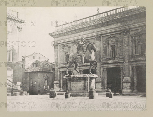 Marcus Aurelius, Capitol - Rome, page, photomechanical print, photographer: Samuel J. Beckett, Piazza del Campidoglio, c. 1890 - c. 1900, Rome, paper, collotype, photoprint, height 152 mm x width 202 mm, sculpture, square, circus, equestrian statue, A grayscale photograph of a city square dominated by a large equestrian statue on a raised stone pedestal, showing a robed rider seated on a rearing horse with one front leg raised, surrounded by short cylindrical bollards and a cobblestone ground; behind the statue stands a grand classical building with tall columns, ornate window frames, and a balustraded roofline with small standing statues, while to the left a smaller building with tiled roof and steps leading up beside a facade with decorative cornices and a window is visible, and a few lamp posts and silhouettes of people appear near the building entrances.