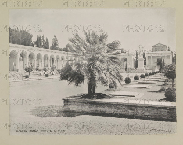 Modern Roman cemetery, page, photomechanical print, photographer: Samuel J. Beckett, Rome, c. 1890 - c. 1900, paper, collotype, photoprint, height 152 mm x width 200 mm, churchyard, cemetery, Verano Cemetery, A wide outdoor scene shows a paved courtyard with a raised rectangular planter in the foreground containing a fan-leaved palm, a row of flat stone slabs set into the ground beside neatly trimmed spherical shrubs, and a low brick retaining wall along the planter edge; to the left a long colonnade with repeated arches and niches runs back toward a stand of tall dark trees, with ornamental plants and monuments beneath the arches; toward the right a tall statue of a robed figure stands on a pedestal behind an ornamental fence, and behind it a classical building with columns and a pediment is visible, framed by additional trees and shrubbery under a pale sky.