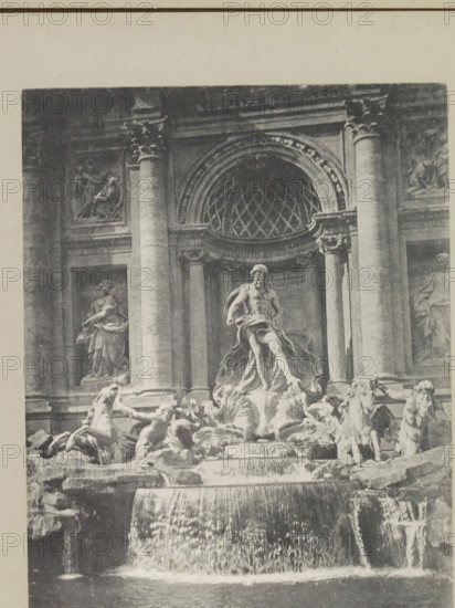 Trevi fountain, page, photomechanical print, photographer: Samuel J. Beckett, Rome, c. 1890 - c. 1900, paper, collotype, photoprint, height 202 mm x width 147 mm, sculpture, (story of) Neptune (Poseidon), ornamental fountain, A grayscale photograph showing a large ornate fountain set against a stone façade with tall fluted columns and decorative relief panels; a central sculpted male figure with draped cloth stands on a rocky plinth surrounded by dynamic sculpted horses and human figures integrated into cascading water features; tiered basins and flowing water spill over carved ledges into a lower pool, while an arched niche with a patterned semicircular grille sits above the central figure and architectural cornices and pilasters frame the scene.