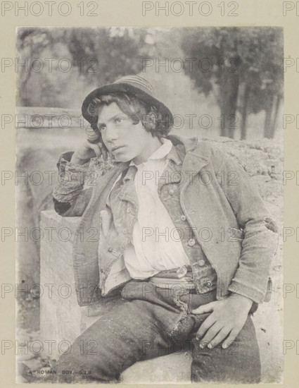 A Roman boy, page, photomechanical print, photographer: Samuel J. Beckett, Rome, c. 1890 - c. 1900, paper, collotype, photoprint, height 202 mm x width 151 mm, youth, adolescent, A black-and-white photograph of a young person wearing a brimmed hat and layered, worn clothing, leaning against a stone surface with one elbow propped and hand resting against the side of the head, eyes looking slightly away from the camera, curly hair visible beneath the hat, a loosely buttoned jacket over a lighter shirt with a visible collar and frayed edges, trousers with a belt and a hand resting on the thigh, a textured stone or brick background and blurred foliage in the upper background.