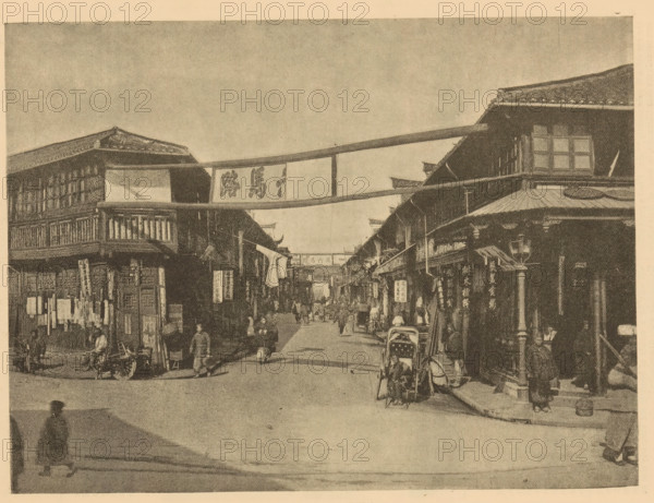 A street in Shanghai, page, photomechanical print, Shanghai, printmaker: Meisenbach's Autotype Company (possibly), c. 1881 - in or before 1886, Meisenbach's Autotype Company, photographer, paper, autotypie, height 157 mm x width 209 mm, street, façade (of house or building), A wide street scene with two- and three-story wooden buildings lining both sides, featuring overhanging roofs, balconies, and hanging banners between the buildings; a covered cart or rickshaw is parked on the right edge and another wheeled cart is visible on the left, with multiple pedestrians walking or standing along the street and clustered near shopfronts; shop signs and vertical hanging placards are attached to building facades, and fabric banners stretch across the street overhead; the image has a grainy, sepia-toned appearance and shows architectural details like tiled roofs, wooden railings, and windowed upper floors.