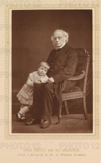 Portrait of John Bright and his grandson, John Bright and his grandson, page, photomechanical print, photographer: A. Whitham, Rochdale, c. 1874 - in or before 1884, printmaker, Manchester, paper, Woodburytype, Woodburytypepie, height 144 mm x width 95 mm, Print after title page., Person, John Bright, grandparent(s) with grandchild(ren) (family group), An older man with white hair and sideburns sits on an ornate wooden chair with carved details and upholstered seat, wearing a dark suit and shoes, while a young child in a light dress, socks, and dark shoes leans against his knee and rests their hands on his leg, both posed against a plain dark backdrop within a decorative rectangular border.