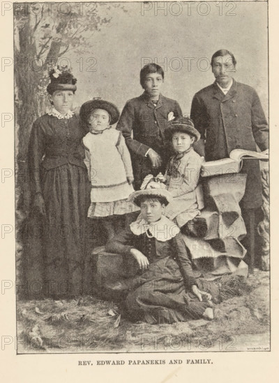 Portrait of Edward Paupanekis and his family, Rev. Edward Papanekis and family, page, photomechanical print, Canada, maker: Waterlow & Sons Limited, c. 1882 - in or before 1892, photographer, North America, paper, autotypie, height 135 mm x width 97 mm, Person, Edward Paupanekis, human races, peoples, nationalities, family, descent, A sepia-toned vintage studio photograph showing a group of people arranged in formal poses; a standing woman on the left wears a dark dress with a high lace collar and a hat decorated with flowers, next to a young girl in a light pinafore and a round hat leaning slightly; a standing boy in a dark jacket with a hand resting on a seated girl's shoulder stands center, the seated girl wearing a dress with a small hat and holding a small object in her lap; a man in a dark coat stands on the right beside a draped table with an open book on it; in front a young woman reclines on the floor wearing a dress with a large white collar and a hat, one arm resting on a ra...