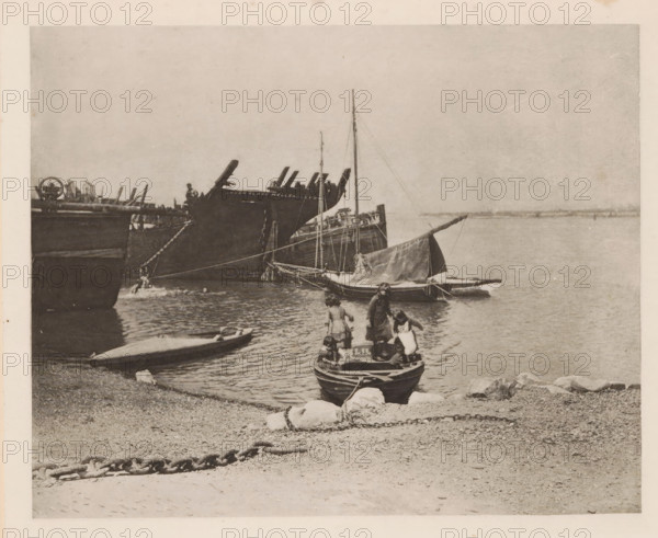 Children in a rowboat on the Breydon, page, photomechanical print, number: 'Plate IV', title: 'Old hulks', photographer: Peter Henry Emerson, Breydon, cliché maker: Walter L. Colls, c. 1880 - in or before 1890, Norfolk (county), paper, photoaquatint, height 161 mm x width 201 mm, rowing-boat, canoe, child, ships (in general), A shoreline scene with small rowboats and larger sailing vessels moored near the water, a small boat pulled up on the pebbled beach, a larger boat in the foreground with people standing and sitting inside it, a sailboat with its sail furled floating nearby, taller ships with masts and rigging visible further out, calm water extending toward the horizon, a chain lying on the beach in the foreground, scattered rocks along the water's edge, and an overcast sky above.