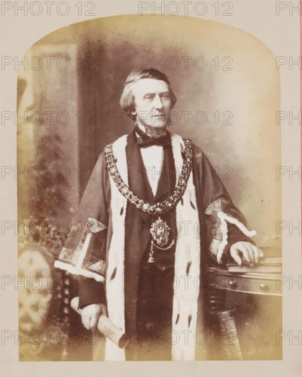 Portrait of Mayor Abel Heywood, photograph, page, photographer: J. McLeod, Manchester, c. 1876 - in or before 1877, paper, albumen print, height 217 mm x width 164 mm, Photo on frontispiece., Person, Abel Heywood, mayor, burgomaster, A sepia-toned portrait of a middle-aged man standing, wearing a dark formal garment with a light fur-trimmed robe draped over his shoulders and ornamental embroidered cuffs, a large decorative chain or collar with an ornate medallion rests around his neck, he has neatly combed hair, sideburns, and a trimmed beard, a bow tie and waistcoat are visible beneath the robe, one hand rests on a wooden table with carved details and metal studs while the other holds a rolled document, the background shows a softly blurred backdrop and patterned fabric at one side, the image has rounded top corners and a faded, aged appearance.