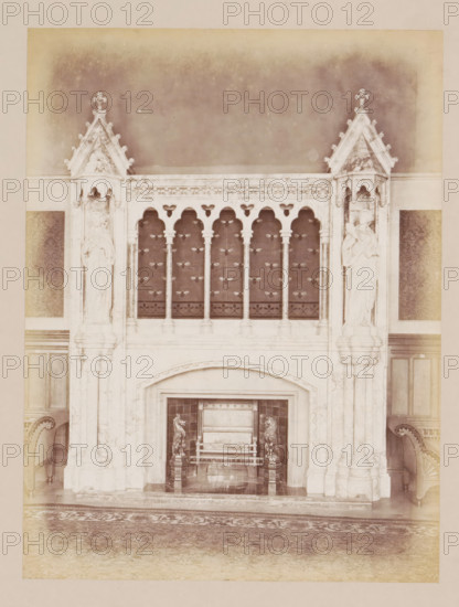 Fireplace in the reception room in Manchester Town Hall, photograph, page, title: 'The Reception-Room Chimney-Piece', photographer: J. McLeod, Manchester Town Hall, 1877, Manchester, paper, albumen print, height 200 mm x width 151 mm, town hall, interior, representation of a building, chimney-piece, mantelpiece, A tall ornate stone fireplace with carved pillars and gothic arches occupies the center of the image, flanked on each side by sculpted figures set into pointed canopied niches; above the fireplace is a decorative balustrade with slender columns and a patterned dark panel behind them, and below the arch of the fireplace opening is an ornate hearth area containing a bench or seat with sculpted animal figures at its sides; portions of upholstered chairs are visible at the left and right edges, and a patterned rug covers the floor in front of the fireplace.