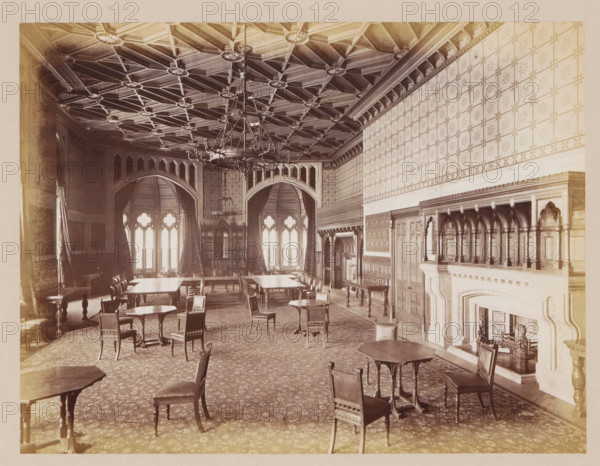 Banquet hall in Manchester Town Hall, photograph, page, title: 'The Banqueting-Room', photographer: J. McLeod, Manchester Town Hall, 1877, Manchester, paper, albumen print, height 162 mm x width 214 mm, celebration meal, feast, banquet, town hall, interior, representation of a building, A large ornate hall with a patterned ceiling featuring geometric woodwork and decorative rosettes, a central chandelier hanging from the ceiling, tall arched windows with tracery and heavy drapery along the far wall, multiple wooden tables and chairs arranged across a patterned carpeted floor, carved wooden paneling and built-in shelving along the walls, an elaborate carved stone fireplace with sculpted details and a decorative overmantel, and a series of archways and columns dividing the space.