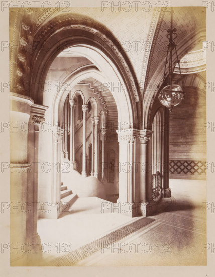 Staircase in Manchester Town Hall, photograph, page, title: 'The Circular Staircases', photographer: J. McLeod, Manchester Town Hall, 1877, Manchester, paper, albumen print, height 213 mm x width 162 mm, townhall, staircase, interior, representation of a building, A sepia-toned interior scene showing a series of ornate stone arches and columns leading to a curved staircase, with multiple slender columns topped by decorative capitals and pointed archways visible through successive openings, a patterned vaulted ceiling with geometric motifs above, a suspended glass lantern hanging from ornate metal chains on the right, carved floral and foliate details on column capitals and arch mouldings, a decorative railing beside the staircase, and a tiled floor with a perforated grate and patterned border in the foreground.