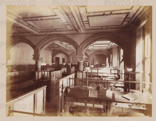 Office in Manchester Town Hall, photograph, page, title: 'The Gas Office', photographer: J. McLeod, Manchester Town Hall, 1877, Manchester, paper, albumen print, height 162 mm x width 213 mm, office (, administration), town hall, interior, representation of a building, A large interior space with high decorative ceilings and multiple arched openings supported by columns with carved capitals, wooden paneling along the walls, rows of wooden desks and partitions forming cubicles, several tall lamps with bowl-shaped shades mounted on stands, a broad wooden table in the foreground with drawers and a closed book resting on it, railings and stair-like platforms dividing levels, tall windows on the right casting light and shadows across the room, and ornamental moldings and recessed panels on the ceiling.