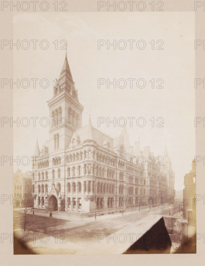 Exterior of Manchester Town Hall, photograph, page, title: 'Cooper Street Front', photographer: J. McLeod, Manchester, 1877, paper, albumen print, height 201 mm x width 150 mm, town hall, façade (of house or building), Manchester Town Hall, A large stone building with ornate Gothic Revival architecture fills the scene, featuring a tall central tower with a steep spire, arched windows, decorative cornices, and multiple smaller turrets and gables; the facade shows rows of tall arched openings and a prominent arched entrance with columns at the corner of a quiet street, where lamp posts line the sidewalks and a single pedestrian stands near the entrance; adjacent streets and neighboring buildings are faintly visible, and the photograph has a pale, faded tone with a wide border and some vignette shadowing at the bottom corners.