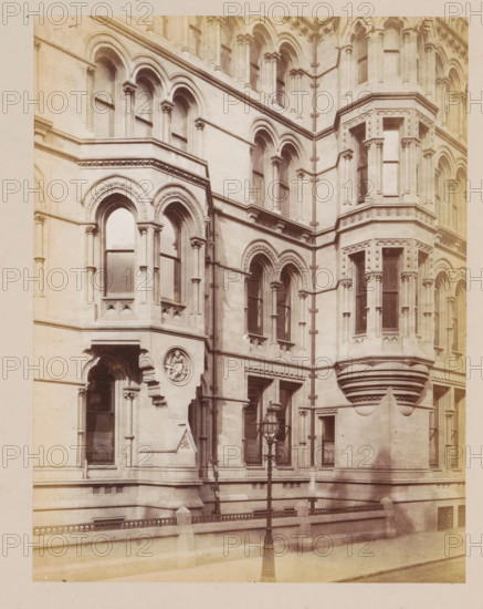 Facade of Manchester City Hall, photograph, page, title: 'The Lloyd Street Façade', photographer: J. McLeod, Manchester, 1877, paper, albumen print, height 213 mm x width 162 mm, town hall, façade (of house or building), Manchester City Hall, A stone building façade with ornate Gothic-style arches and columns, featuring tall arched windows set within recessed frames and decorative moldings, a projecting octagonal bay with narrow vertical windows and layered cornices, a round carved medallion inset into the wall, a recessed doorway beneath an arch, a cast-iron street lamp mounted on a slender post in front of the building, a low stone balustrade with decorative metal railing along the sidewalk, and smooth stone pavement at the base.