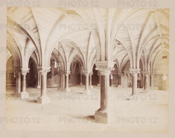 Sculpture hall in Manchester City Hall, photograph, page, title: 'The sculpture hall', photographer: J. McLeod, Manchester City Hall, 1877, Manchester, paper, albumen print, height 162 mm x width 212 mm, townhall, A large interior space with a series of stone columns topped by ornate capitals supporting ribbed, vaulted arches that form repeated pointed curves across the ceiling, a smooth pale floor with subtle rectangular patterns, carved column bases resting on square plinths, decorative moldings and recessed archways along the perimeter, a suspended metal chandelier hanging near a stair with a carved balustrade, a checkerboard patterned panel set into one wall, and a few sculptural busts or reliefs mounted on pillars and walls.