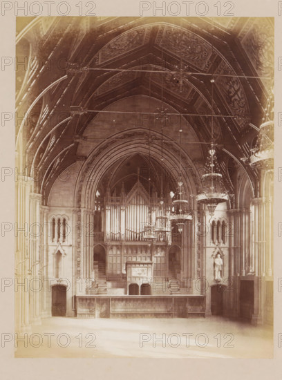 Vestibule in Manchester City Hall, photograph, page, title: 'The Public Hall', photographer: J. McLeod, Manchester City Hall, 1877, Manchester, paper, albumen print, height 202 mm x width 152 mm, vestibule, hall, town hall, A high vaulted interior with ornate ribbed arches and decorative ceiling panels, large hanging chandeliers suspended by rods, a tall pipe organ with visible vertical pipes set within an elaborate wooden case, stone columns and carved architectural details flanking the space, steps leading up to the organ platform, niches with statues, arched doorways at ground level, a long wooden balustrade or shelving unit along the front, and soft light filtering into the large hall.