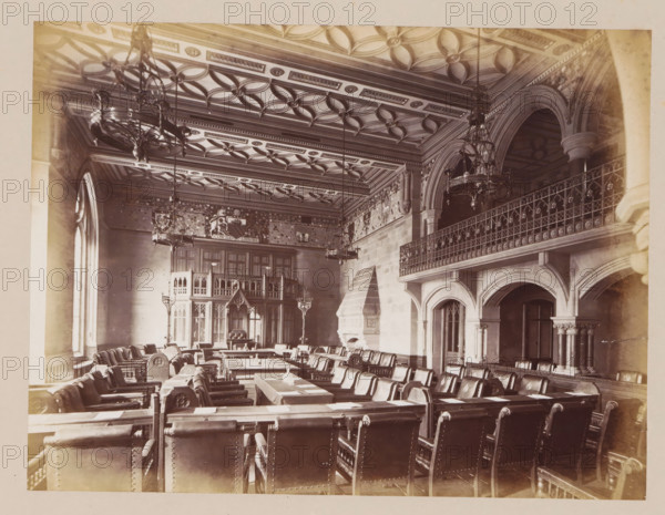 Meeting room in Manchester City Hall, photograph, page, title: 'The Council Chamber', photographer: J. McLeod, Manchester City Hall, 1877, Manchester, paper, albumen print, height 162 mm x width 204 mm, townhall, interior, representation of a building, provincial government, A large ornate hall with a patterned coffered ceiling and multiple hanging chandeliers, rows of leather-upholstered chairs arranged around long wooden tables, tall arched windows along one wall, a carved stone fireplace with a stepped hood, a wooden carved screen or gallery at the far end, decorative wall panels and friezes above the paneling, an elevated balcony with an intricate metal railing supported by stone arches and columns, and various lamps and candlesticks placed throughout the room.
