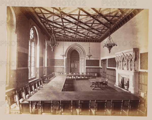 Mayor's Parlour in Manchester City Hall, photograph, page, title: 'The Mayor's Parlour', photographer: J. McLeod, Manchester City Hall, 1877, Manchester, paper, albumen print, height 161 mm x width 212 mm, town hall, interior, representation of a building, A large rectangular room with a patterned coffered ceiling featuring diagonal beams and decorative panels, three ornate hanging chandeliers suspended from the ceiling, tall arched windows along one long wall with framed panels beneath them, long wooden tables arranged in a U shape surrounded by high-backed wooden chairs, an arched doorway with layered stone molding centered on the far wall, dark wooden wainscoting and wall panels lining the room, an elaborately carved stone fireplace with clustered columns and decorative arches on the right wall, a smaller standalone table and chairs near the fireplace, and a patterned floor covering the central area.