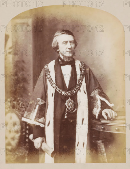 Manchester town hall, book, inscription: 'In commemoration, Hall Sub-Committee.', inscription: 'ex libris SFJ', photographer: J. McLeod, 1877, plant material, biological material, paper, wood (plant material), leather, printing, albumen print, color on cut, height 330 mm x width 275 mm x thickness 42 mm, A sepia-toned portrait of a middle-aged man standing and facing forward, wearing a dark formal coat with a high collar and a bow tie, layered with a fur-trimmed ceremonial robe and an ornate chain of office draped around his shoulders; his hair is parted to one side and he has distinctive sideburns and a short beard on his chin, his left hand rests on a wooden table with carved details while his right hand holds a rolled document, and the background shows a softly blurred interior with mottled tonal variations.