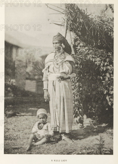 Woman with a child at Kullu, Himachal Pradesh, India, page, photomechanical print, caption: 'A KULU LADY', photographer: Frederick Saint John Gore, Kullu, c. 1890 - in or before 1895, printmaker, Kullu Valley, paper, collotype, photoprint, height 179 mm x width 129 mm, adult woman, child, ornaments, jewels, A sepia-toned photograph of a standing woman and a seated child outdoors, the woman wearing a headwrap, layered necklaces and facial jewelry, a shawl draped over her shoulders and a long patterned skirt, her hands resting at her waist with one holding an object, and the child seated on the ground in front of her wearing a cap and simple clothing; foliage and a thatched structure appear in the background.