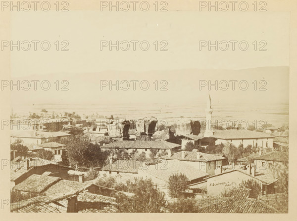 View of Alasehir, photograph, page, photographer: Alexander Svoboda, Alasehir, c. 1859 - in or before 1869, Ottoman Empire, paper, albumen print, height 128 mm x width 177 mm, Photo for page 35., ruin of a building, architecture, A sepia-toned panoramic view of a town with clustered tiled rooftops in the foreground, scattered trees and vegetation among the buildings, a mosque with a slender minaret rising above the structures toward the right, several ruined stone arches or large rock formations near the center, low-lying fields stretching into the distance, and a faint silhouette of a mountain ridge along the horizon under a pale sky.