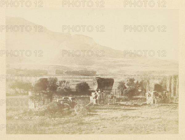 View of the ruins of a church in Sardis, photograph, page, photographer: Alexander Svoboda, Sardis, c. 1859 - in or before 1869, Asia Minor, paper, albumen print, height 128 mm x width 177 mm, ruin of church, monastery, A faded landscape photograph showing a hilly backdrop with a sloping ridge and plateau, a middle ground of rocky terraces and scattered low stone ruins, and a foreground of grassy fields with stone blocks and partially standing stacked stone structures interspersed with small trees and shrubs.