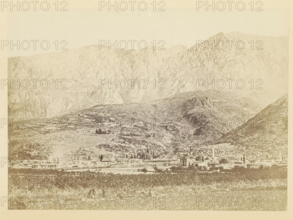 View of the ruins of Magnesia on the Sipylos, photograph, page, photographer: Alexander Svoboda, Asia Minor, c. 1859 - in or before 1869, paper, albumen print, height 129 mm x width 179 mm, ruin of a building, architecture, (high) hill, Magnesia on the Sipylos, A wide landscape photograph showing a mountainous backdrop with steep rocky slopes and ridges, a midground of rolling hills with scattered trees and terraced areas, and a foreground containing a flat field with rows of vegetation and a few small groups of people standing or sitting; along the base of the hills there is a spread of low buildings and a taller tower-like structure among them, and the overall image has a faded, sepia-toned appearance.