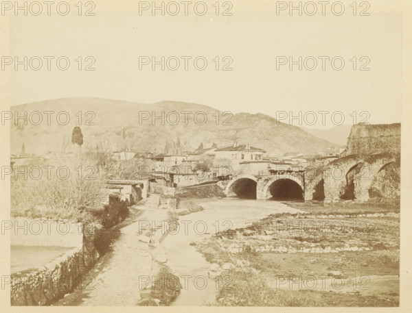 View of Pergamon, photograph, page, photographer: Alexander Svoboda, Pergamon, c. 1859 - in or before 1869, Asia Minor, paper, albumen print, height 124 mm x width 176 mm, city-view in general, 'veduta', bridge in city across river, canal, A sepia-toned landscape showing a river running toward the foreground with a dirt path alongside stone walls, a stone arched bridge crossing the river with multiple arches, adjoining stone structures and low buildings clustered near the bridge, a taller building with a tiled roof near the center, leafless shrubs and trees along the banks, a large rounded hill rising in the background, and expanses of sky above.