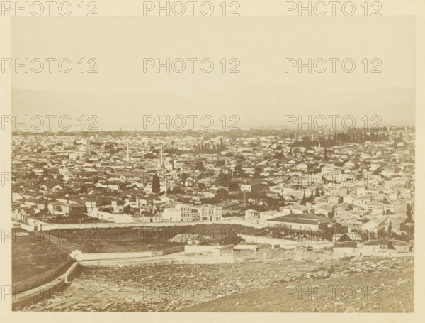 View of Smyrna, photograph, page, photographer: Alexander Svoboda, Smyrna, c. 1859 - in or before 1869, Ottoman Empire, paper, albumen print, height 130 mm x width 178 mm, city-view in general, 'veduta', A wide sepia-toned panoramic view showing an expansive cityscape with densely packed low-rise buildings and varied rooflines, several slender vertical towers rising among the structures, clusters of tall trees interspersed through the urban area, a low stone wall running across the foreground above a textured, rocky slope, and a pale sky occupying the upper portion of the image.