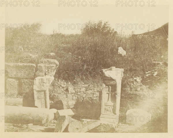 Ruin of the tomb of Saint Luke at Ephesus, photograph, page, photographer: Alexander Svoboda, Ephesus, c. 1859 - in or before 1869, paper, albumen print, height 142 mm x width 181 mm, ruin of a building, architecture, grave, tomb, A faded photograph showing a grassy, overgrown hillside with scattered stone ruins in the foreground, including carved stone blocks and upright rectangular fragments with recessed panels and relief detailing, broken cylindrical columns lying on the ground, rough stacked stone walls, uneven rocky terrain, and dense vegetation covering much of the slope.
