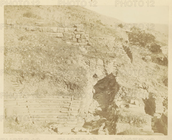 The Odeon of Ephesus, photograph, page, photographer: Alexander Svoboda, Ephesus, c. 1859 - in or before 1869, Efeze, paper, albumen print, height 139 mm x width 177 mm, ruin of a building, architecture, A sepia-toned scene showing a rocky hillside with irregular stone steps carved into the slope and a rough stone retaining wall partially visible, scattered rocks and sparse grasses covering much of the terrain, a narrow dark crevice or opening in the rock face, and low vegetation and small shrubs dotting the hillside.