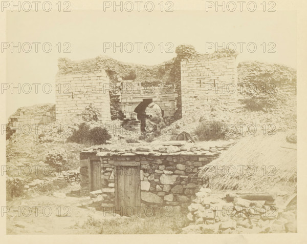 View of the Gate of Persecution on the hill of Ayasoluk, photograph, page, photographer: Alexander Svoboda, Asia Minor, c. 1859 - in or before 1869, paper, albumen print, height 134 mm x width 174 mm, gate, entrance, A sepia-toned photograph showing stone ruins on uneven ground with a partially collapsed arched gateway set between two tall masonry walls, rubble and low stone structures in front of the gateway, a small stone building with a wooden door in the foreground, scattered rocks and vegetation around the site, and a thatched roof visible at the lower right edge.