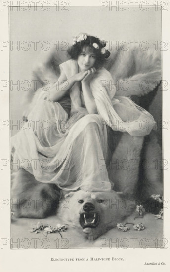 Portrait of an unknown woman on a polar bear skin, page, photomechanical print, printmaker: Lascelles & Co., c. 1889 - in or before 1899, photographer, paper, autotype, electrotype from a half-tone block, height 176 mm x width 111 mm, adult woman, beasts of prey, predatory animals: polar bear, A seated young woman with a wreath of flowers in her hair rests her chin on her hands, wearing a flowing translucent gown that drapes around her and cascades over a large fur beneath her; behind her the fur forms a voluminous backdrop, and at her feet a bear skin with an open mouth and visible teeth lies flat on the ground, with scattered petals and small flowers near the bear’s head.
