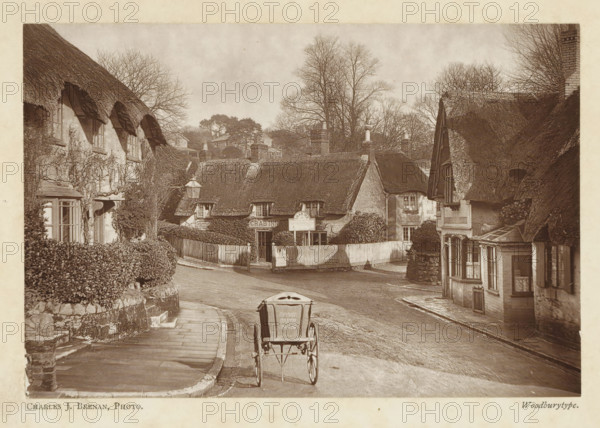 Crab Inn, Shanklin, Isle of Wight, page, photomechanical print, photographer: Charles J. Brenan, c. 1889 - in or before 1899, printmaker, paper, Woodburytype, Woodburytypepie, height 98 mm x width 145 mm, inn, coffee-house, public house, street, A sepia-toned street scene shows a narrow road curving past thatched cottages with low, textured roofs and small multi-paned windows, flanked by hedges and stone walls. A simple two-wheeled cart is positioned on the road in the foreground, viewed from behind. On the left, a paved footpath runs beside a stone wall topped by trimmed bushes and leads toward a row of cottages partly covered in climbing plants. In the center, a fenced entrance opens to a cluster of cottages with a lamp post nearby and chimney stacks rising above the thatch. On the right, another thatched building presents a projecting bay window and timbered details, with bare trees visible behind the rooftops and additional houses set further back on a gentle rise. The sky appears ...