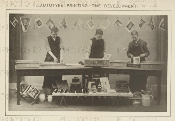 Process of developing what is believed to be a carbon print demonstrated by three men, Autotype Printing - The development, page, photomechanical print, inscription: 'I. Mounting (use of the Squeegee). 2. Developing Prints. 3. Fixing in Alum Solution. A. Cold Water Tray, No. IB Blotting Boards. C. Mounting Dish and Stool. D. Developing Tank. E. Cold Water Tray No. 2. F. Alum Bath G. Cold Water Tray No. 3. H. 'Hero' Stove. J. Johnson's Actinometers. R. Burton's Actinometers. S. Sawyer's Actinometers. T. Pressure Frames. P. Autotype Pressure Boxes. V. Packets of Cut Tissues. W. Waxing Compound. X. Collodion.', printmaker: Sawyer, printer (person): Autotype Company, The, c. 1885 - in or before 1895, Sawyer, Autotype Company, photographer, paper, collotype, photoprint, height 107 mm x width 175 mm, Print on a fold-out sheet opposite page 38., developing, film, A sepia photograph shows three men standing behind a long wooden table, each wearing dark aprons over shirts with rolled sleeves an...