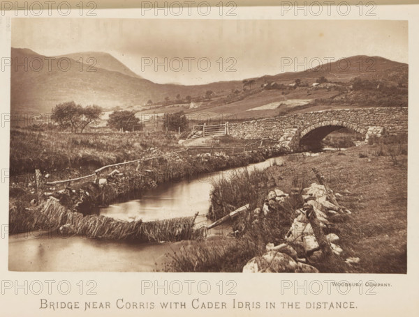 Bridge near Corris with Cader Idris in the distance, Bridge in a landscape near Corris, with a view of Cadair Idris in the distance, page, photomechanical print, Wales, printmaker: Woodbury Permanent Photographic Printing Company, c. 1881 - in or before 1891, photographer, paper, Woodburygravure, height 90 mm x width 141 mm, mountains, bridge, A sepia-toned landscape photograph showing a gently flowing stream bordered by grassy banks and scattered stones, a rustic wooden fence running alongside the water, a stone arch bridge crossing the stream, low rolling hills and terraced fields in the background, clusters of trees dotting the midground, and distant rounded mountains under a cloudy sky.