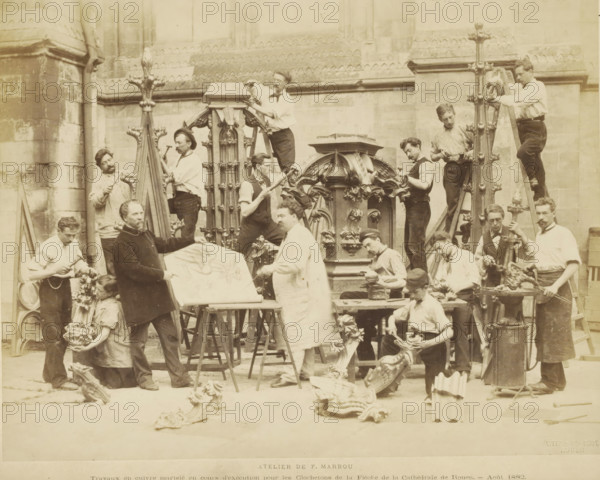 Atelier de F. Marrou Travaux en cuivre tortureé en cours d'exécution pur les Clochetons de la Flèche de la Cathédrale de Rouen., Sculptors at work for the studio of Ferdinand Marrou at Notre-Dame in Rouen, photograph, page, name: 'Witz & cie Phot. Rouen', photographer: Witz et Cie., Rouen, 1882-08, Witz et Cie., photographer, paper, albumen print, albumendruk, height 214 mm x width 273 mm, piece of sculpture, reproduction of a piece of sculpture, sculptor at work, A sepia-toned photograph shows a group of men and a woman working on ornate carved and cast decorative architectural pieces outside a stone building with buttresses and patterned masonry; several tall vertical carved finials and a central elaborate canopy-like structure are prominent, with men standing on ladders or holding tools while others shape or polish smaller decorative elements on tables and the ground; a few workers wear aprons or smocks and hold hammers, files, or clamps, while one man points at a large flat panel d...